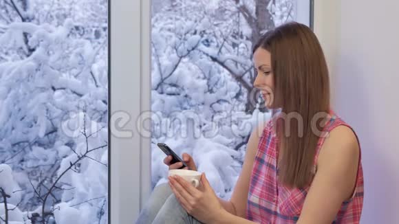 漂亮的女孩坐在窗台上用智能手机喝咖啡冬天在外面视频的预览图