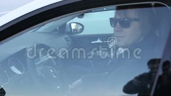迷人的年轻人坐在汽车的车轮后面戴着黑色眼镜说话视频的预览图