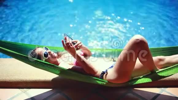 美丽的年轻苗条的比基尼女郎躺在吊床上在美丽的阳光下在游泳池里打电话视频的预览图