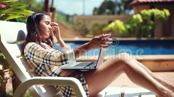 年轻的黑发女人在她的笔记本电脑上工作躺在池边的日光浴床上漂亮的女模特自由职业视频的预览图