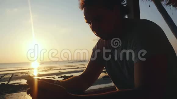 在海滩上的酒吧休息的帅哥在美丽的夕阳下使用手机发送信息慢动作视频的预览图