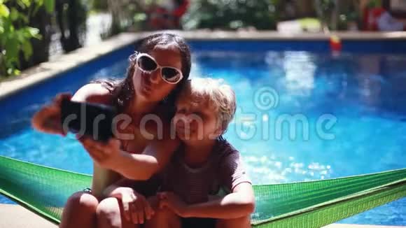 年轻的黑发女人和她的儿子坐在吊床上用手机在游泳池里自拍家庭幸福假期快乐视频的预览图