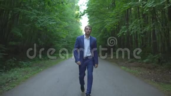 穿着蓝色西装的年轻商人正在森林里沿着马路散步视频的预览图