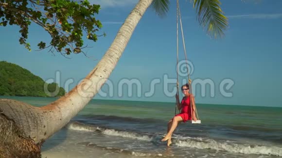 美丽的女孩在海滩的秋千上放松在热带海洋岸边穿着红色连衣裙的女人穿上的女人视频的预览图