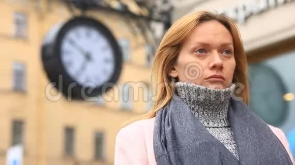 一个穿着粉红色外套和毛衣的女人站在时钟附近拥挤的街道中间等待着视频的预览图