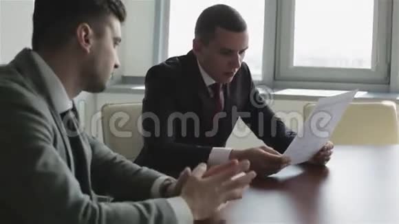 两个穿西装的商人正坐在窗前的一张桌子旁学习一份新合同视频的预览图