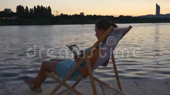 女性坐在海滨的日光浴床上阅读电子阅读器视频的预览图