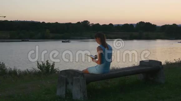 年轻女子在公园日落时看一本电子书视频的预览图