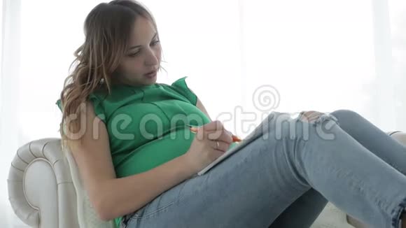 年轻的孕妇穿着便服坐在大窗户附近的沙发上写日记视频的预览图