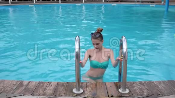 穿比基尼的苗条女人在蓝水背景下的梯子上从游泳池里爬出来年轻的女人视频的预览图