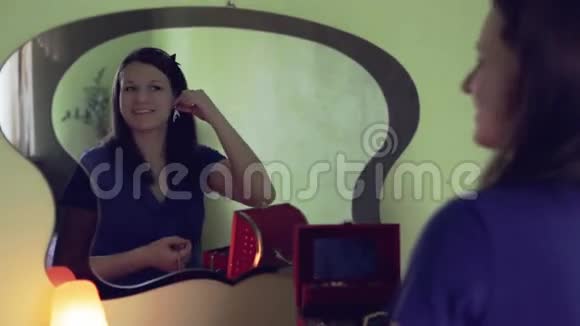 年轻漂亮的女孩穿着蓝色连衣裙坐在镜子前试穿耳环视频的预览图