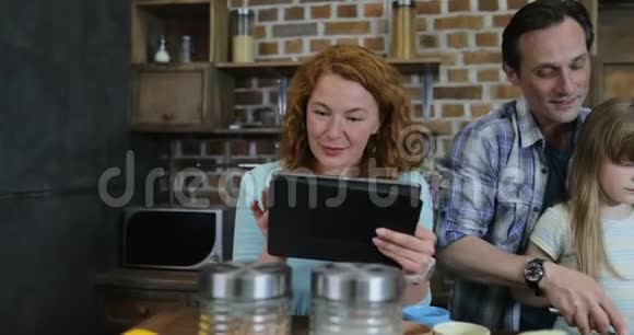 快乐的家庭在厨房烹饪父亲和女儿切碎蔬菜而母亲在网上阅读食谱视频的预览图