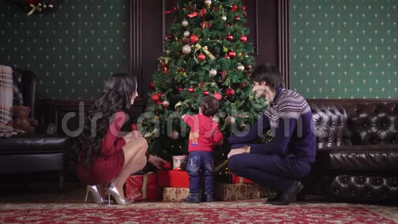 年轻的一家人坐在圣诞树附近它是装饰花环玩具丝带蝴蝶结妈妈爸爸和他们的孩子视频的预览图