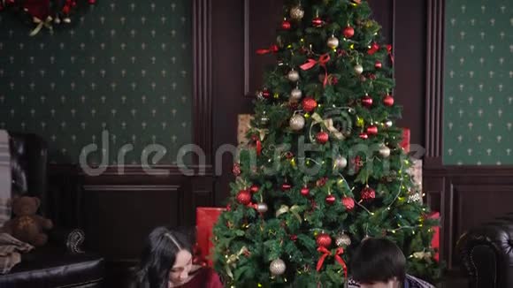 穿着考究的幸福家庭坐在圣诞树附近的地毯上在温暖的客厅里爸爸妈妈视频的预览图