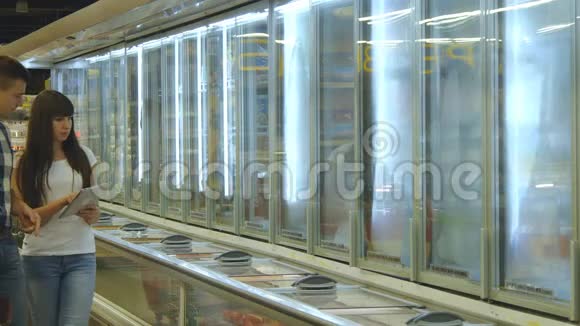 这对年轻夫妇正站在杂货店的冰箱旁用冷冻食品挑选包装从中获取产品的人视频的预览图