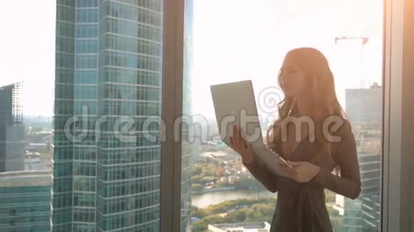 年轻的美女靠近窗户在城市背景上使用笔记本电脑视频的预览图
