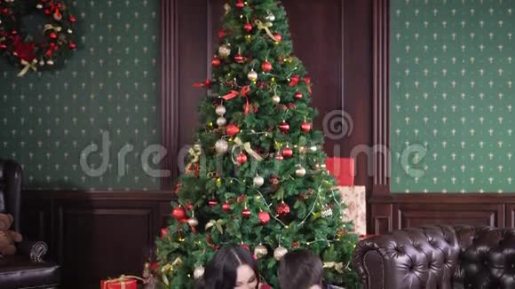 一个年轻幸福的家庭坐在一棵装饰精美的圣诞树旁开始读一本他们年幼儿子的书视频的预览图