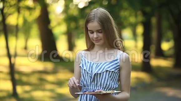 美丽的金发女孩坐在凳子上用颜料和铲子在公园里画一幅画视频的预览图