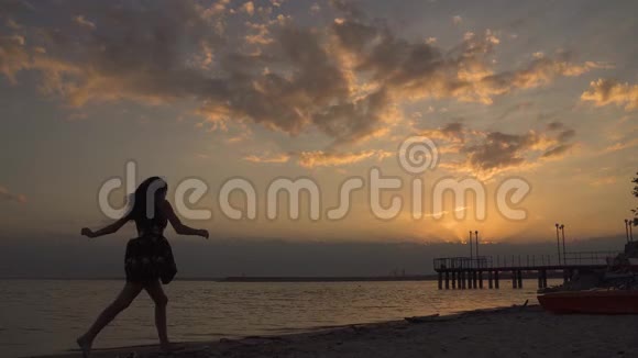 夕阳下海滩上一个穿着浅色连衣裙的女孩的剪影美丽的小女孩光着脚在沙滩上奔跑视频的预览图