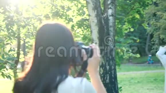 一位穿着白色衬衫的漂亮女孩职业摄影师正在一个公园里用柔软的衣服拍一个快乐的微笑女孩的照片视频的预览图