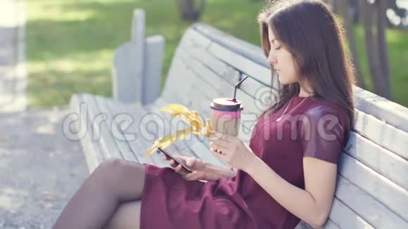 一个美丽快乐的年轻女孩外表迷人手里拿着手机坐在城市公园的长凳上视频的预览图