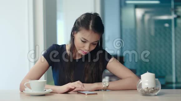 迷人的年轻女子一边喝咖啡一边在咖啡馆里看手机视频的预览图