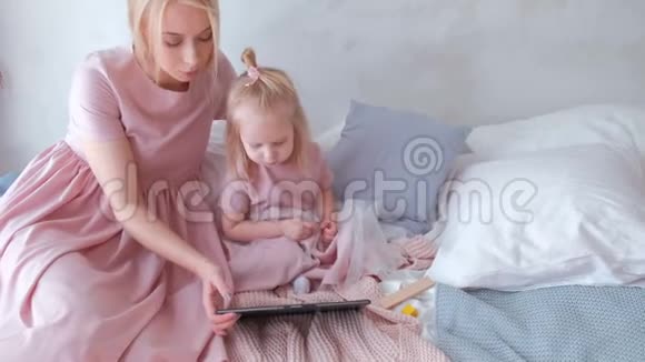 年轻迷人的金发女人和她的小迷人的女儿穿着粉红色的连衣裙看着平板电脑里的东西视频的预览图