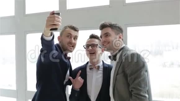 三个有代表性的英俊男人穿着西装和蝴蝶结在大窗户附近自拍视频的预览图