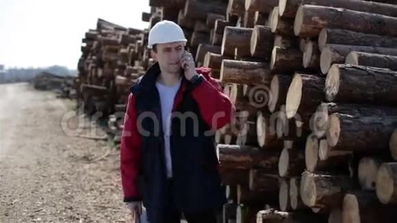 一个戴着白色头盔和红色夹克的建筑工人在堆叠的原木附近的手机上交谈视频的预览图