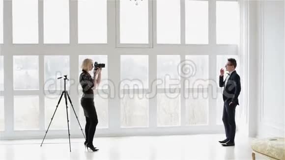 一个穿黑色裙子的金发女孩正在大窗户附近拍摄一个穿西装的男人的照片视频的预览图