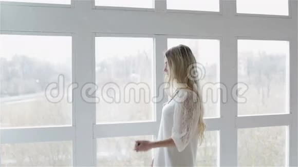 一个穿着白色时尚连衣裙的女孩正在手机上输入号码并在窗户附近交谈视频的预览图