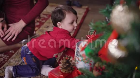 小男孩在地毯上玩并试图得到一个盒子与礼物躺在圣诞树下孩子非常视频的预览图
