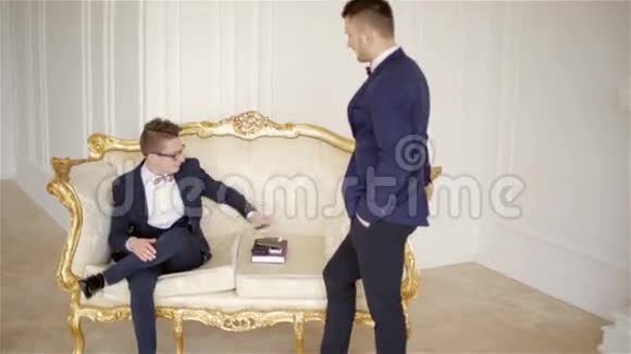 两个穿着蝴蝶结西装的有代表性的男人在一个漂亮的白色房间里和优雅的金色沙发交流视频的预览图