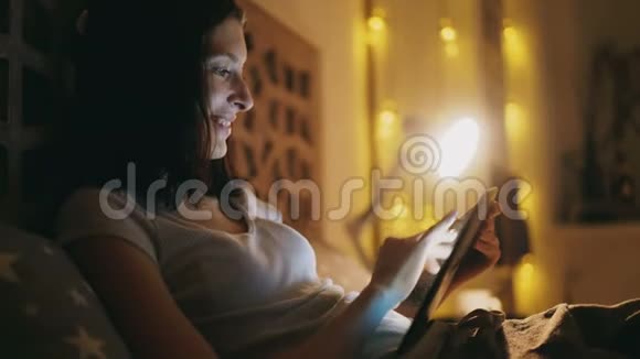 快乐微笑的女人用平板电脑分享社交媒体晚上躺在家里的床上视频的预览图