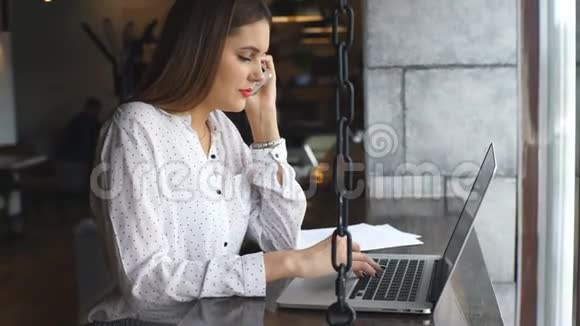 一位年轻漂亮的女士她拿着笔记本电脑和智能手机与坐在窗边的顾客交谈视频的预览图