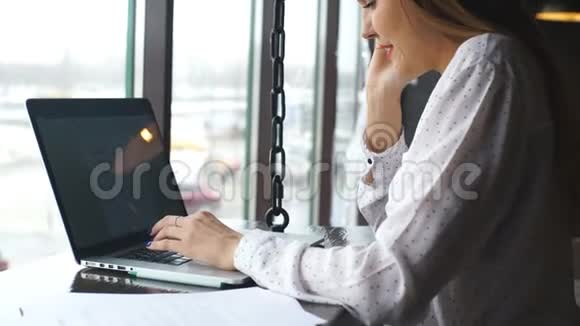 一位年轻漂亮的女士她拿着笔记本电脑和智能手机与坐在窗边的顾客交谈视频的预览图