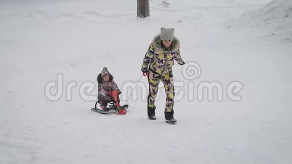 年轻的母亲把女儿卷在公园里的雪橇上冬季活跃家庭休闲可爱的女人和女孩一起玩视频的预览图