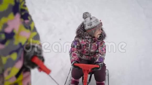 母亲骑着一个小女孩坐在雪橇上方向盘在公园的雪道上快乐的孩子在看着周围视频的预览图