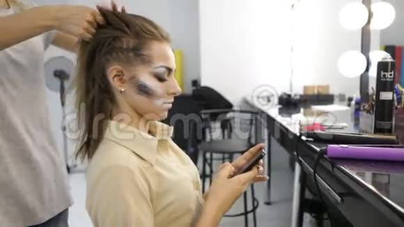 女孩在时尚工作室使用智能手机而理发师则在客户的头发上工作视频的预览图
