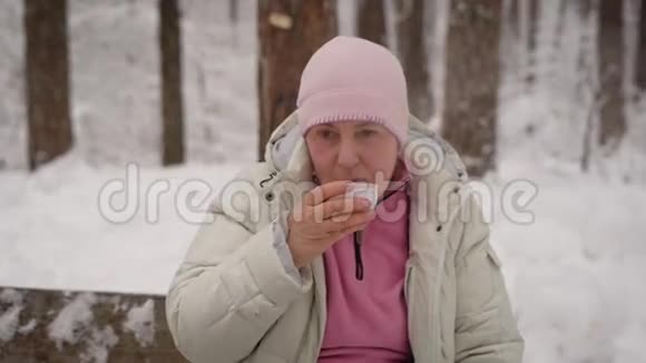 在白雪覆盖的公园里一位老妇人坐在长凳上喝着热茶祖母在新鲜的艾里散步后视频的预览图