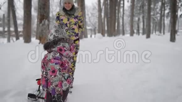 小女孩独自拉着雪橇穿过公园里的雪年轻的母亲在后台观看和帮助视频的预览图