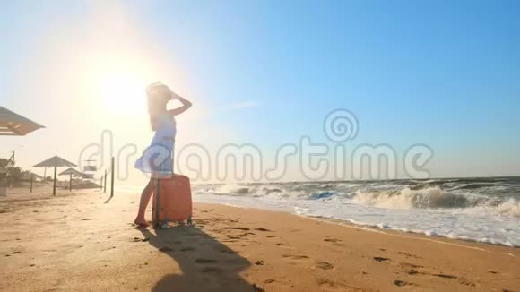 一个年轻的女人带着一个手提箱坐在海滩上一个年轻的女孩带着一个轮式手提箱沿着海滩散步一路走来视频的预览图