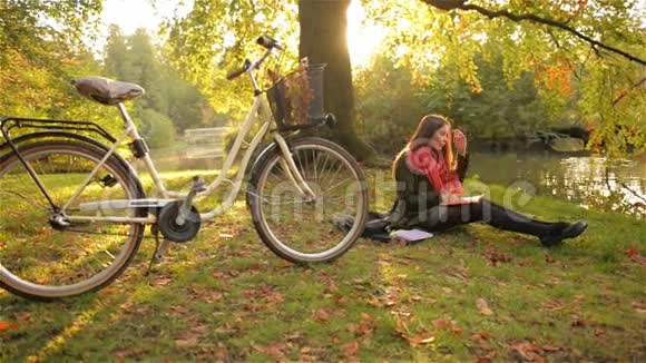 快乐的学生女孩放松在草写在笔记本上自行车和美丽的公共公园的背景视频的预览图