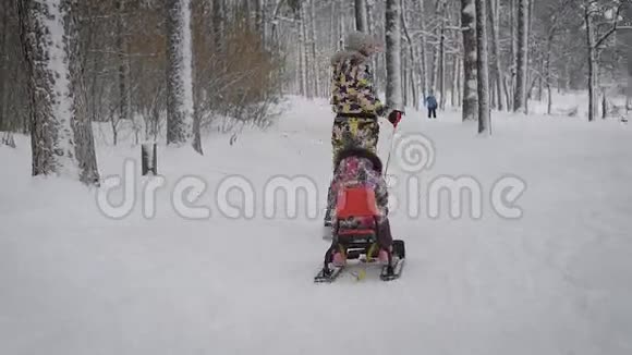 年轻快乐的母亲在冬天的森林里的小路上和她的小女儿一起沿着红色的雪橇前行视频的预览图