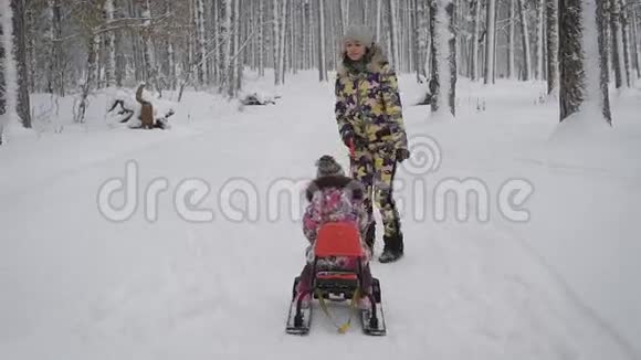 年轻的妈妈在雪橇上翻滚一个小女孩他们在一个冬天的公园里玩耍那里长着很多高大的树两个女孩视频的预览图