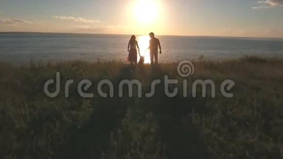 快乐的爸爸妈妈站在悬崖边上把女儿的双手举海边和夕阳下视频的预览图