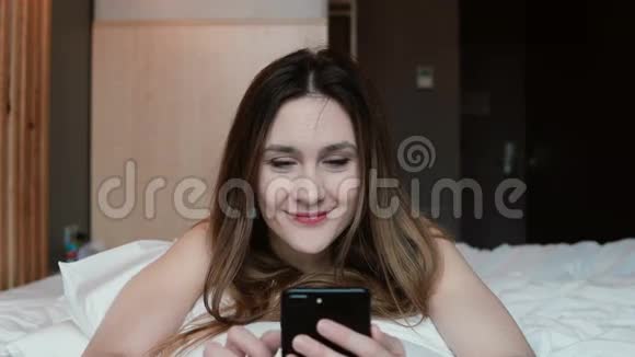 年轻的女人躺在床上用智能手机女孩上网和人聊天微笑视频的预览图