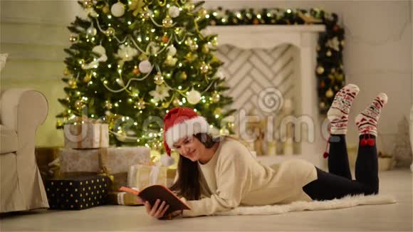 美丽的布鲁内特与圣诞帽阅读一些有趣的故事躺在地板上圣诞装饰的家女伴视频的预览图
