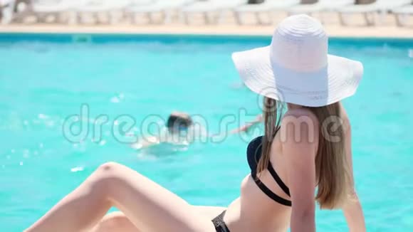 快乐的年轻女孩穿着黑色泳衣和白色帽子坐在室外游泳池的边缘把脚伸进水里视频的预览图