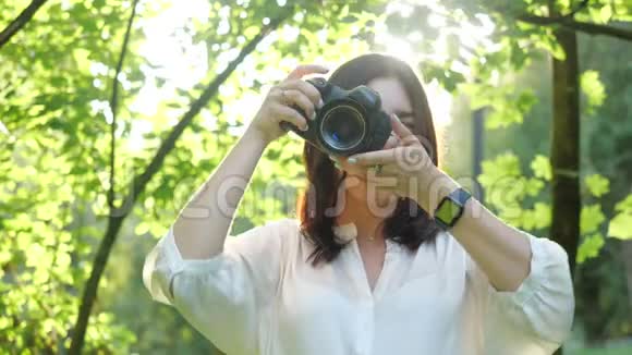 美丽的微笑女孩摄影师穿着白色衬衫正在公园里拍摄绿色树叶的柔和背景视频的预览图